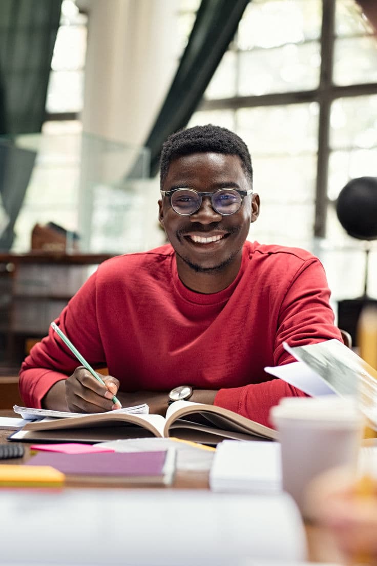 Étudiant noir souriant avec lunettes écrivant dans un cahier dans une bibliothèque ensoleillée.